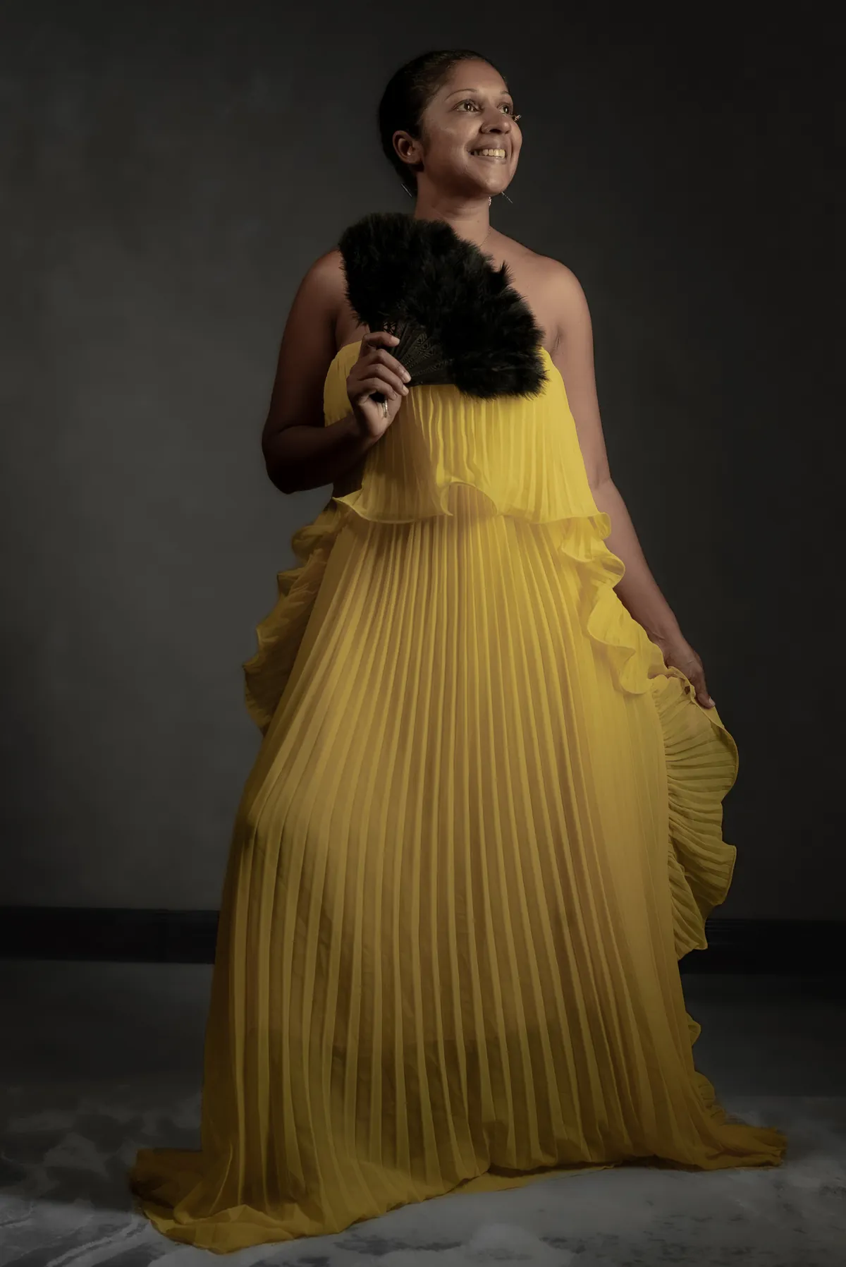 Full-length creative studio photograph of artist Natacha Boodhoo wearing a yellow dress and holding a hand fan