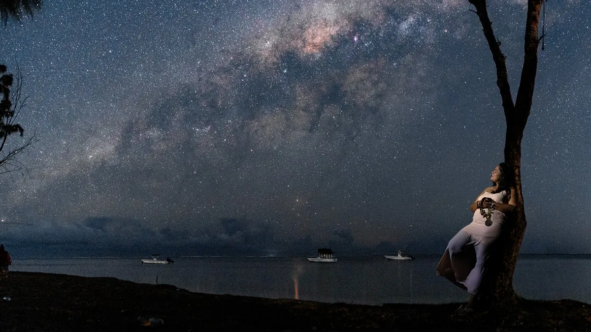 Woman posing beside a tree beneath the Milky Way during a night portrait session by the coast